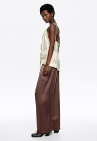 Top de satén beige con detalles de encaje, combinado con pantalones anchos de satén marrón y zapatos de tacón bloque negro, sobre un fondo liso.
