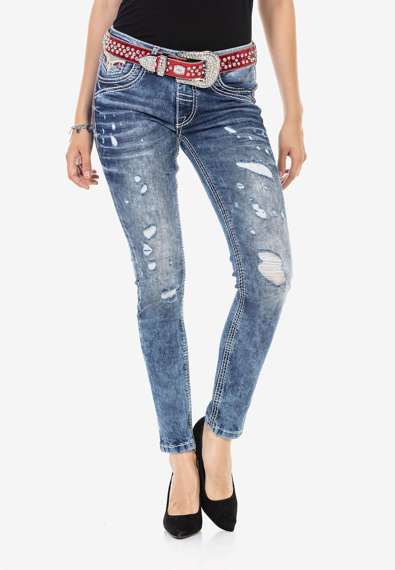 Blaue, ausgewaschene Skinny-Jeans mit ausgefransten Details, versehen mit weißen Nähten und einem roten Nieten-Gürtel. Getragen mit schwarzen High-Heels.