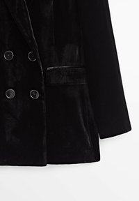 Blazer en velours noir à double boutonnage avec quatre boutons sur le devant, une poche à rabat sur le côté droit et une manche longue sur le côté gauche.