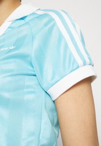 Ljusblå Adidas sporttröja med vita ränder på ärmarna och vit krage, bärs på bar arm mot en plain bakgrund.