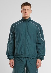 Teal, zip-up jakke med høy krage. Har hvite aksentlinjer og elastiske mansjetter. Laget av et lett, glatt materiale.