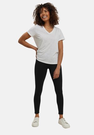 Glunderende vrouw met krullend haar, gekleed in een wit V-hals T-shirt, zwarte legging en witte sneakers, staand met één hand op haar heup.