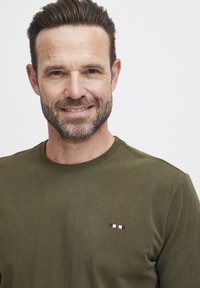 Olíva zöld pamut póló, rövid ujjú, kerek nyakkivágással, a mellrészen egy kicsi hímzett folttal, amely piros, fehér és kék csíkokat ábrázol.