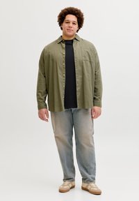 Άνδρας με σγουρά μαλλιά που φοράει πουκάμισο χακί με κουμπιά, μαύρη μπλούζα, ελαφρώς μπλε τζιν και μπεζ αθλητικά παπούτσια, στέκεται μπροστά σε λευκό φόντο.