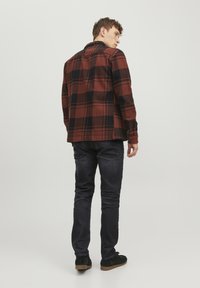 Camisa de franela de cuadros rojos y negros en una persona, vista desde atrás. Combinada con jeans oscuros y zapatos negros. Textura suave, puños abotonados.