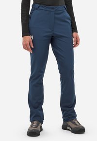 Osoba w granatowych spodniach outdoorowych z kieszenią na zamek błyskawiczny i szarych butach trekkingowych, stojąca na białym tle.