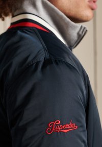 Giacca navy con colletto grigio. Presenta dettagli a righe rosse e bianche e il logo "Superdry" ricamato sulla spalla. Materiale liscio e leggero.
