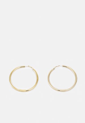 Paire de boucles d'oreilles créoles dorées de taille moyenne avec "GUESS" gravé sur de petites plaques rectangulaires plates près des fermoirs, présentée sur fond blanc.