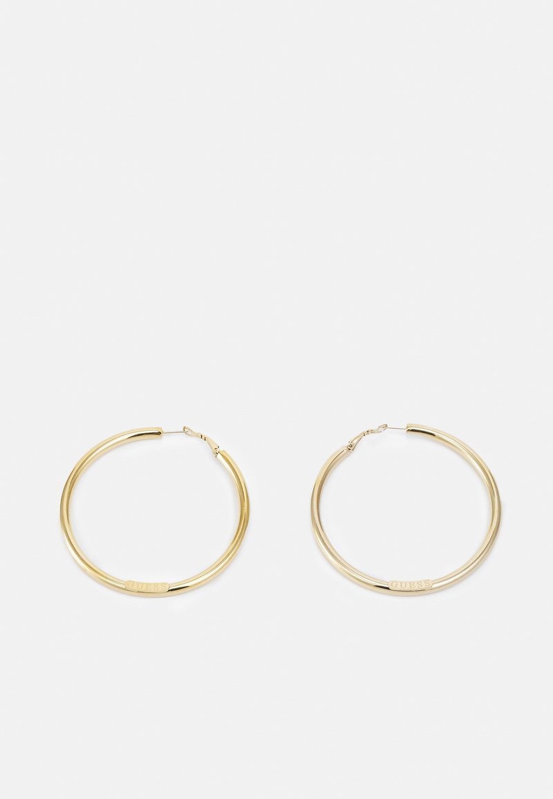 Paire de boucles d'oreilles créoles dorées de taille moyenne avec "GUESS" gravé sur de petites plaques rectangulaires plates près des fermoirs, présentée sur fond blanc.