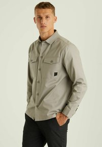 Šviesiai pilka marškinėlių su dviem krūtinės kišenėmis, klasikiniu apykakle ir akcentu logotipo laikikliu. Lygi tekstūra, ilgomis rankovėmis, pritaikyta forma.