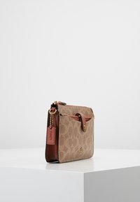 Brązowa torba z materiału z wzorem logo, skórzanymi akcentami, złotymi elementami metalowymi, zamknięciem na klapkę i zawieszką. Kompaktowy, prostokątny kształt.