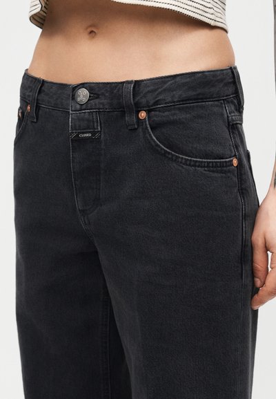 Jean en denim noir de hauteur moyenne porté par une personne portant un haut rayé court, montrant les poches avant et les détails de la fermeture par bouton.