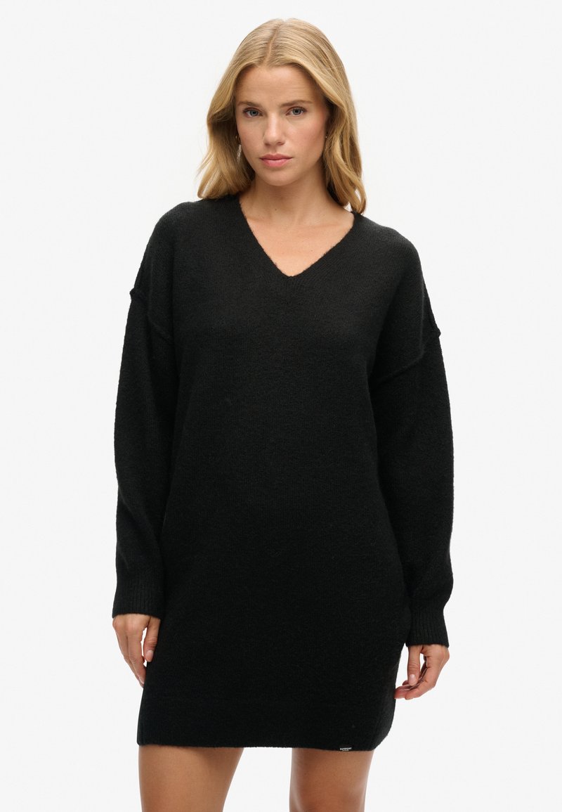 Vestido de punto negro de manga larga con escote en V, ajuste holgado y textura suave. Presenta puños acanalados y un dobladillo ligeramente alargado.