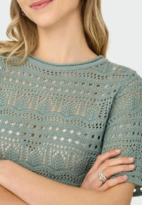 Suéter de punto verde claro con patrón calado, que cuenta con mangas cortas y cuello redondo. Detalle texturizado notable y un acabado suave.