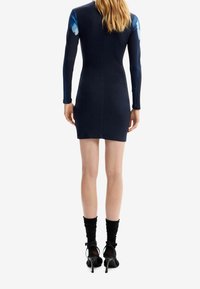 Robe marine à manches longues côtelées avec imprimé papillon bleu sur les épaules ; silhouette ajustée ; portée avec des talons noirs à lanières et des chaussettes montantes jusqu'aux genoux.