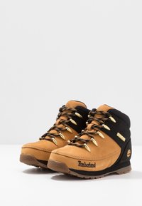 Botas de senderismo de gamuza marrón y tela negra con herrajes dorados, que presentan una suela texturizada y un logo de la marca en el lateral.
