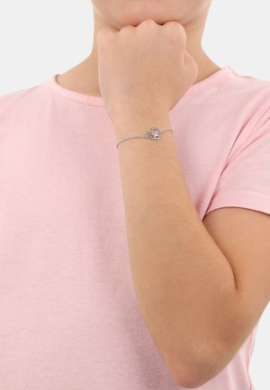 Srebrna bransoletka łańcuszkowa z sercowym zawieszką o różowo-białym wzorze, noszona na nadgarstku w zestawieniu z jasnoróżową koszulą.