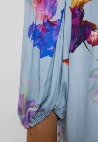 Light blue blouse with puffed sleeve design, featuring large, colorful floral patterns in purple, pink, and yellow. Soft, flowing fabric.