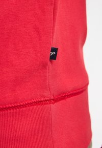 Hanorac roșu din bumbac cu amestec, având textură ribbed, cusături flatlock și un mic etichetă neagră a brandului pe cusătură.