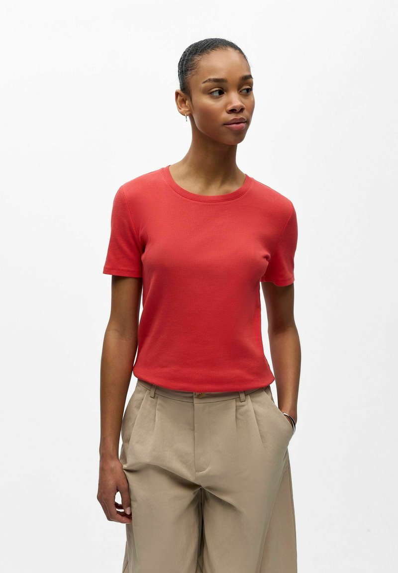Femme à la peau foncée et aux cheveux attachés en chignon, portant un t-shirt rouge à manches courtes et un pantalon beige taille haute, debout devant un fond blanc uni.