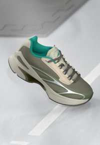 Zapatillas deportivas en tonos verdes y beige con cordones grises y forro interior en color teal, colocadas sobre una superficie gris claro con líneas blancas.