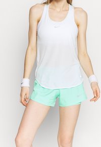 Camiseta deportiva sin mangas blanca con un dobladillo curvado y pequeño logo de Nike, combinada con pantalones cortos verdes claros y muñequeras blancas.