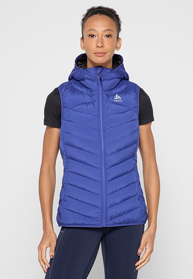 Eine Frau trägt eine blaue gesteppte ärmellose Kapuzenweste, die zugezippt ist, dazu ein schwarzes T-Shirt mit kurzen Ärmeln und marineblaue Leggings. Sie steht vor einem grauen Hintergrund.