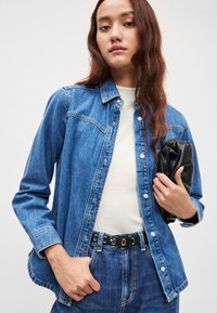 Τζιν μπουφάν σε ανοιχτό μπλε με κούμπωμα με κουμπιά, τσέπες στο στήθος και μακριά μανίκια· συνδυασμένο με λευκή ριμπ μπλούζα και μαύρο κλατς.