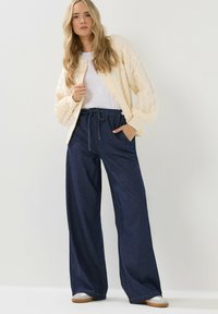 Cremfarbener Kabelstrick-Cardigan, weißes T-Shirt darunter und lockere, dunkle Jeanshose mit elastischem Bund und Taschen. Lässige Schuhe runden das Outfit ab.