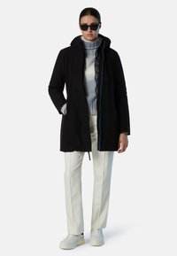 Cappotto nero con cappuccio, tasche frontali e dettagli blu, indossato sopra un dolcevita grigio e pantaloni bianchi a gamba dritta; abbinato a sneakers bianche.