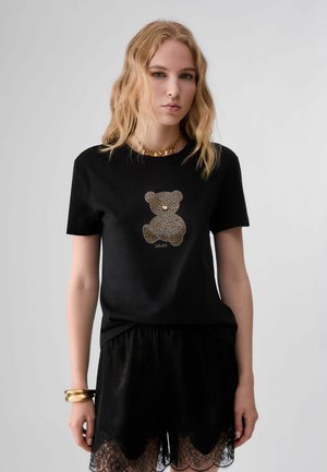 Junge Frau mit welligem blondem Haar trägt ein schwarzes T-Shirt mit goldfarbenem Teddybär-Design und schwarze Shorts mit Spitzenbesatz vor einem schlichten Hintergrund.