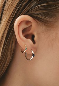 Se muestran tres aros: uno dorado con una piedra verde, uno plateado con una piedra azul y uno plateado con un acento de piedra clara.