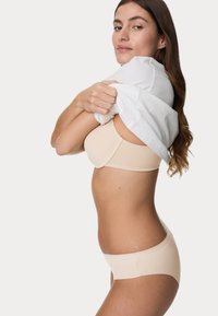 Un reggiseno beige senza cuciture e biancheria intima coordinata su una persona, che mette in mostra un tessuto liscio, un design aderente e uno stile minimalista su uno sfondo neutro.