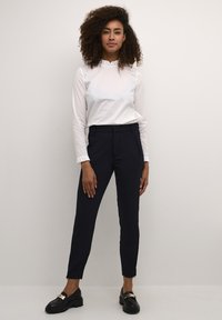 White long-sleeve blouse with ruffled collar paired with dark blue tailored pants. Black loafers with gold accents complete the outfit.