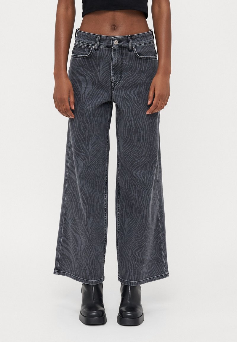 Personne portant un jean taille haute à jambes larges avec imprimé zèbre foncé et des bottines noires, debout devant un fond uni.