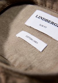 Deux étiquettes blanches sur tissu beige portent les mentions "LINDBERG SLIM FIT" et "COTTON LINEN."