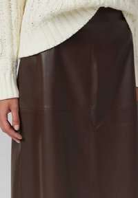 Jupe en cuir brun au toucher lisse, avec des coutures décoratives et une taille haute. Associée à un pull en maille blanche.