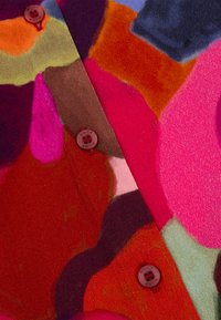 Multicoulore stof met abstracte vormen in tinten van rood, roze, bruin, groen en blauw. Drie rode knopen sieren het shirt.