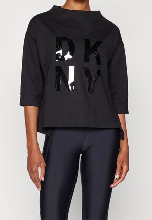 Kvinde iført en sort oversized sweatshirt med skinnende sorte og hvide "DKNY" bogstaver og sorte tætsiddende leggings.