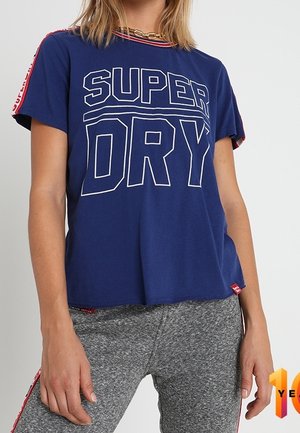 Blauw katoenen T-shirt met witte "SUPER DRY" tekst, korte mouwen en rode accenten op de schouder. Gecombineerd met grijze sportieve broek.
