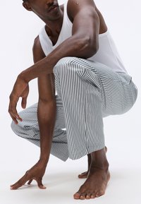 Vīrietis baltā bezpiedurkņu krekla un pelēkām svītrainām biksēm ir nolaidies uz ceļiem basām kājām, ar vienu roku pieskaroties grīdai uz baltā fona.