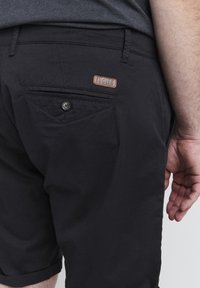 Pantalones cortos negros con un ajuste estructurado, que cuentan con un bolsillo trasero y una pequeña etiqueta de marca marrón. La tela parece suave con un ligero brillo.