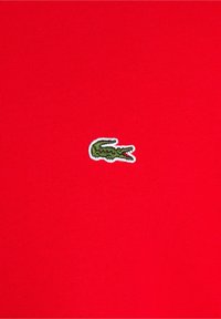Polo rouge présentant un petit logo crocodile vert brodé sur le côté gauche, fabriqué dans un tissu lisse et léger.
