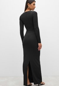 Abito nero a maniche lunghe con un design aderente, dotato di uno spacco sul retro, realizzato in tessuto liscio. Dettagli includono una scollatura bassa sul retro.