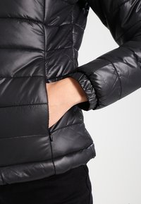 Zwarte gewatteerde jas van glanzend materiaal, met een ritssluiting aan de zijkant en geribbelde manchetten. Horizontale stikdetails zichtbaar.