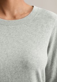 Jasnoszary sweter z prążkowanym dekoltem, gładką teksturą i luźnym krojem, z naciskiem na detale kołnierza i górnej części materiału.