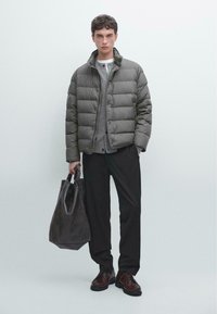 Pufferjacke in Dunkelgrün mit hohem Kragen, grauer Pullover darunter, schwarze Hose, Lederbag und braune Schuhe. Glattes Material und gestepptes Design.