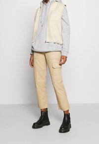 Beżowe spodnie cargo z czterema kieszeniami, szara bluza z kapturem i kremowa kamizelka z polaru. Czarna para botków za kostkę z masywną podeszwą. Prosta, teksturowana warstwowość.