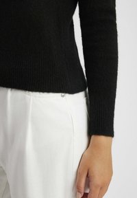 Pull noir en tricot texturé avec une silhouette ajustée et des poignets côtelés, associé à un pantalon blanc présentant un détail de bouton-pression visible.