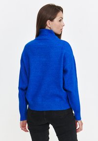 Niebieski, oversize sweter z wysokim kołnierzem, wykonany z miękkiej, teksturowanej dzianiny, z prążkowanym wykończeniem u dołu i na mankietach, stylizowany z czarnymi spodniami.